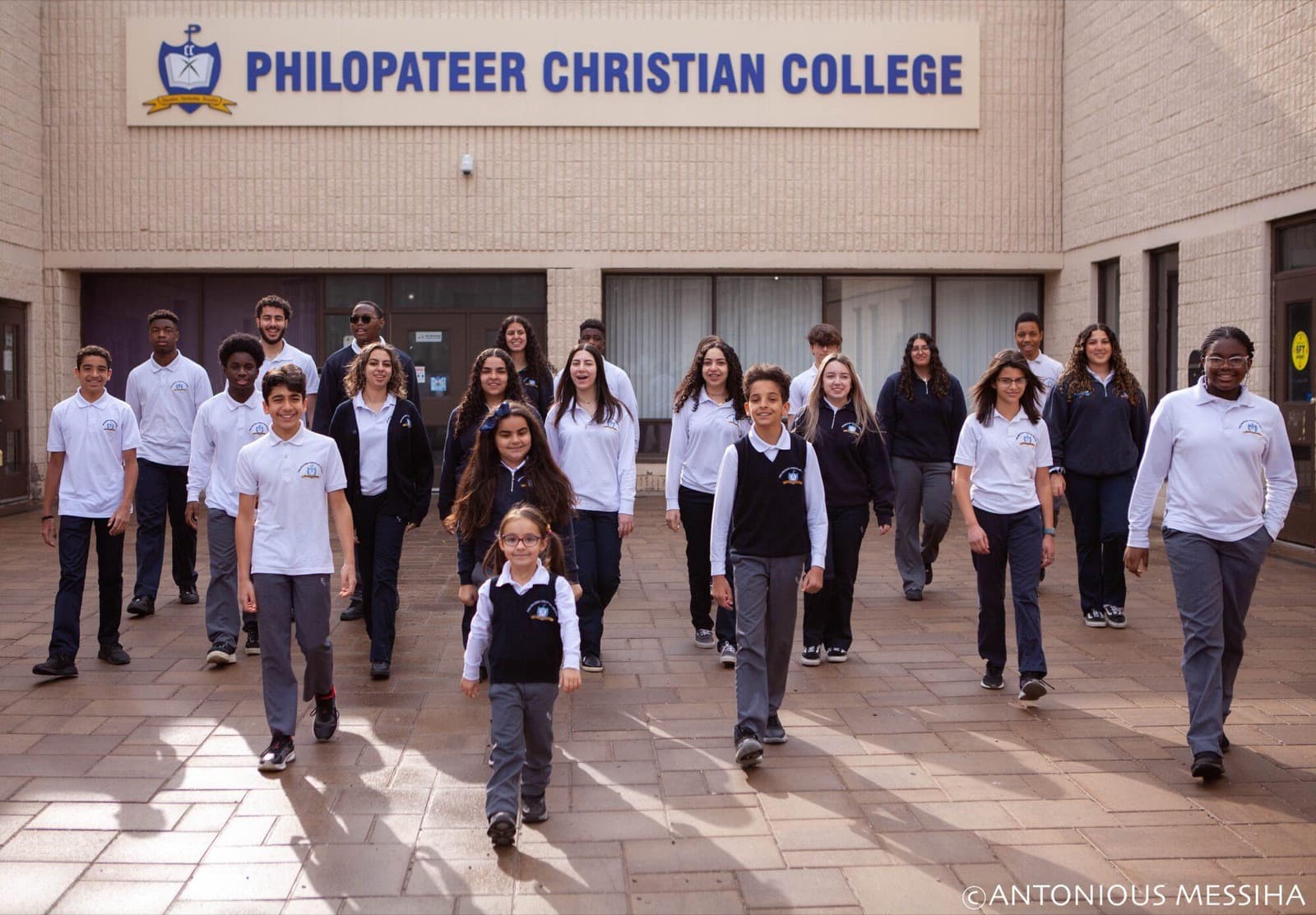 Students in uniform walk across the school courtyard toward the camera, beneath the Philopateer Christian College sign. Photo credit: Antonious Messiha.