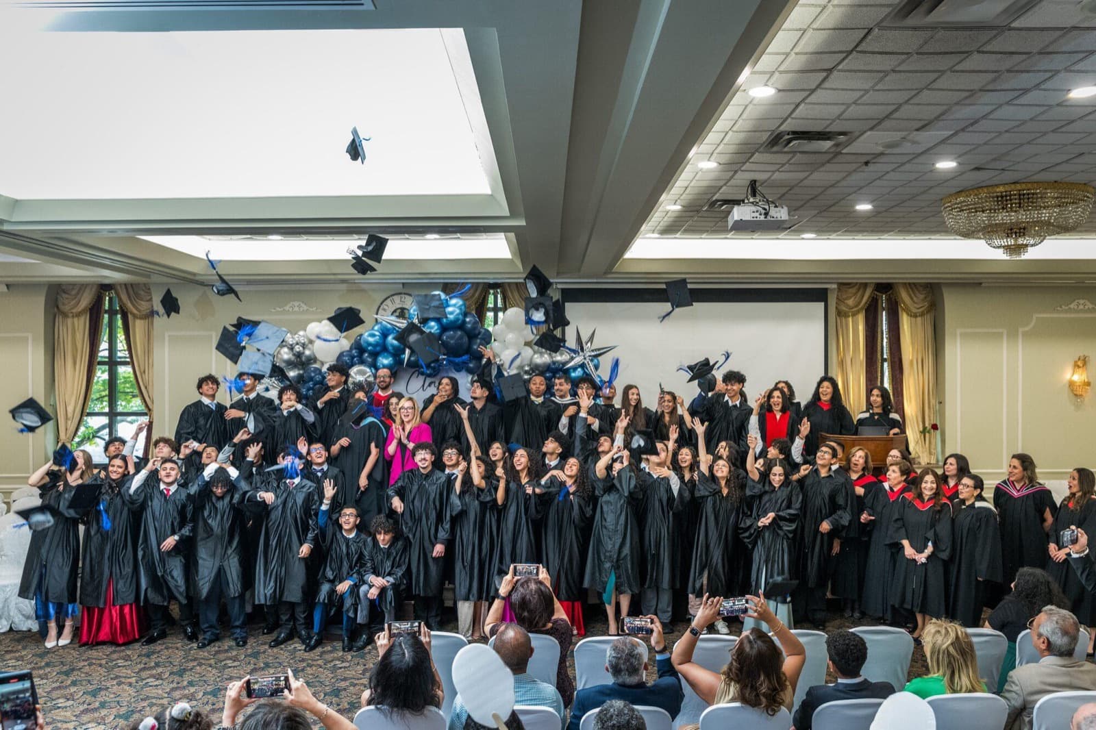 Graduates in gowns throw their caps into the air in a hall decorated with blue and white balloons.