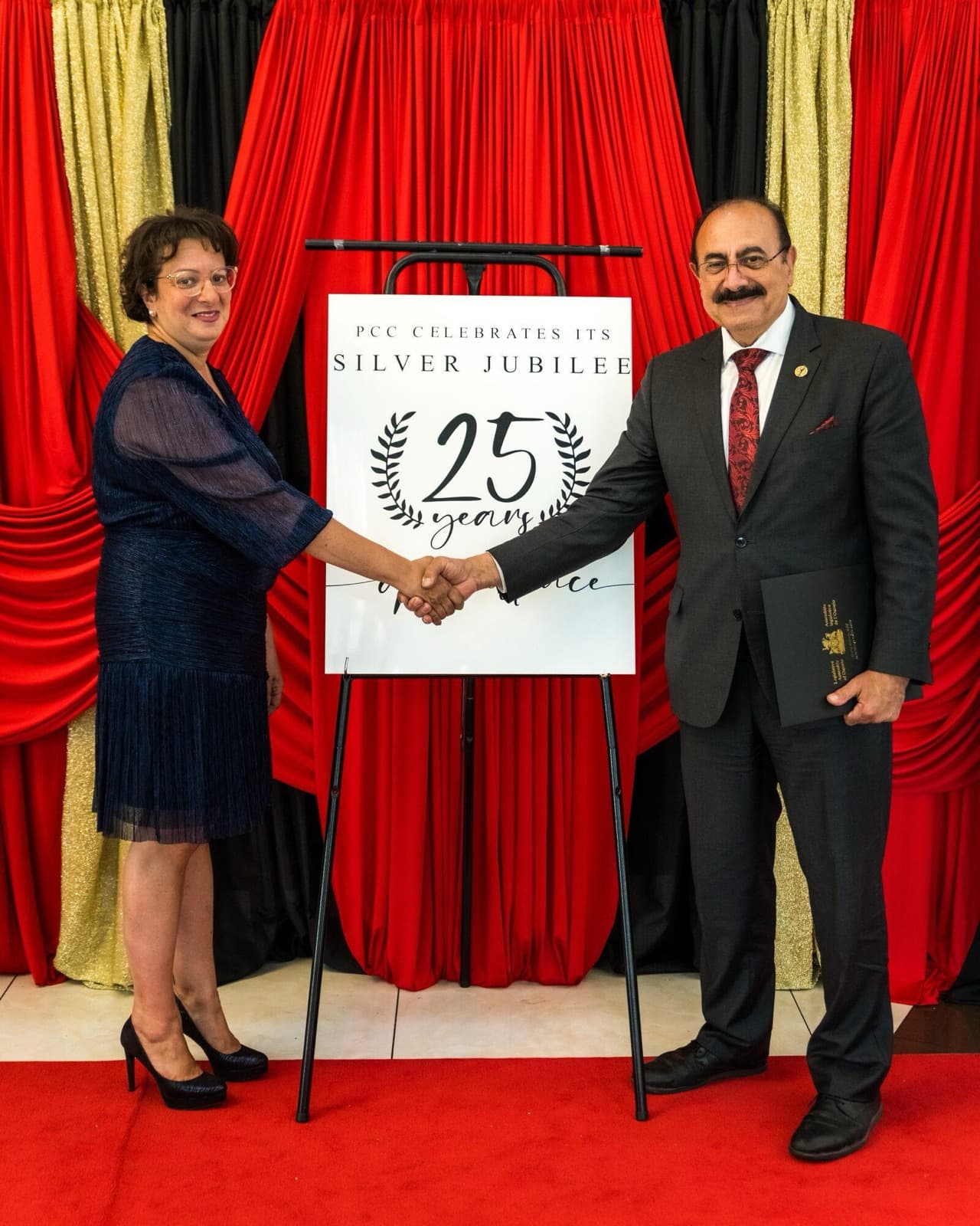Two adults shake hands in front of red curtains and a sign celebrating the school's Silver Jubilee, 25 years.
