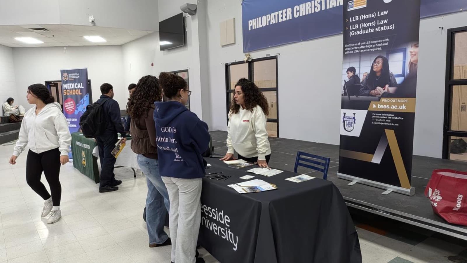 Representatives stand at booths with university banners while students and families walk through the school at a university fair.