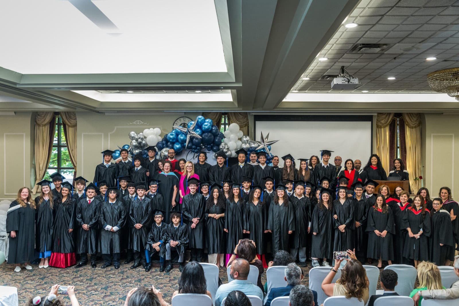 The graduating class stands together in a banquet hall, with family seated in front of them.